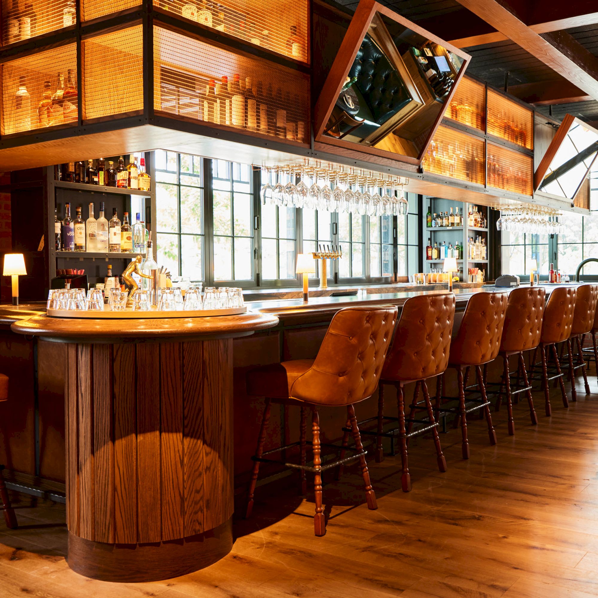 The image shows a modern bar with wooden stools, a long counter, liquor bottles, and glassware under warm lighting in an inviting atmosphere.