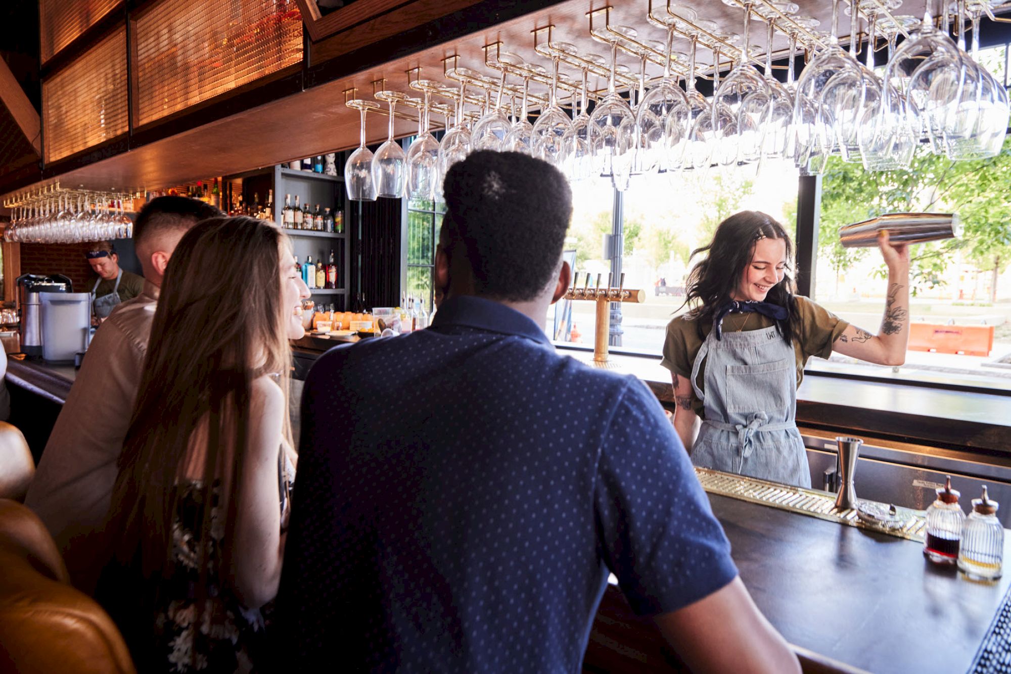 A group of people sits at a bar while a smiling bartender serves drinks, with glasses hanging overhead and bright light outside.