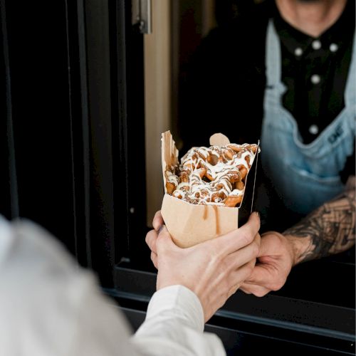 A person in an apron hands a paper container of food, possibly waffles with sauce, to another person through a window.