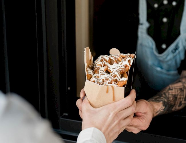A person in an apron hands a paper container of food, possibly waffles with sauce, to another person through a window.