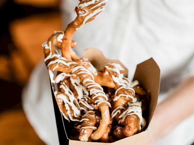 A person holds a box of twisted fries drizzled with a creamy sauce, grabbing one fry.