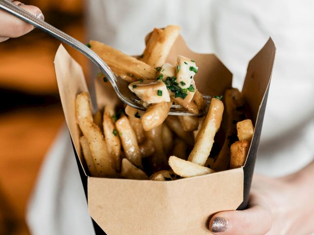 A person is holding a container of fries topped with a creamy sauce and herbs, using a fork to pick some up.