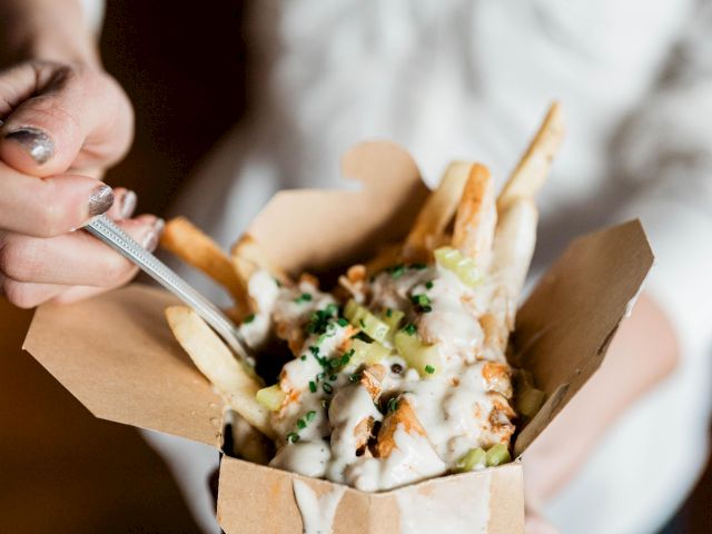 A person holding a box of loaded fries topped with sauce, cheese, and herbs, while using a fork to take a bite.