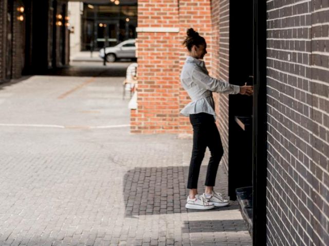 A person is standing on a brick pavement, facing a wall with their hand on a door handle, in an urban setting with parked cars.