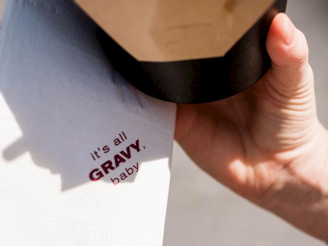 A hand holds a container of fries with gravy, accompanied by a napkin that has the words "it's all GRAVY, baby" printed on it.