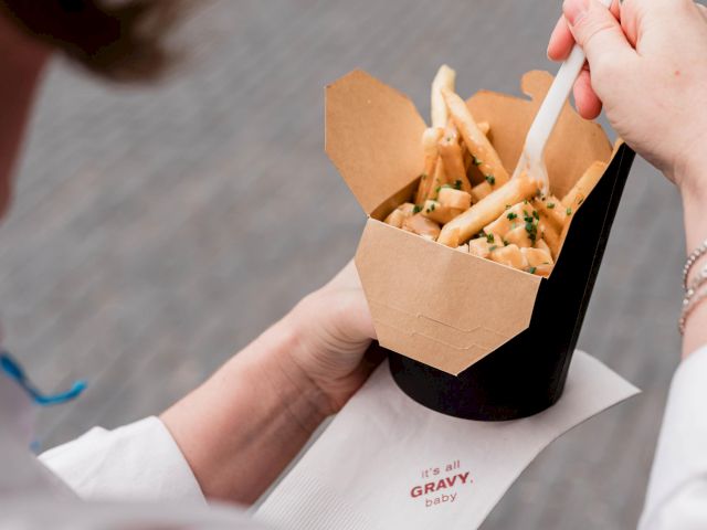 A person is holding a box of fries with a fork, standing on a street or outdoor area, seen from above.