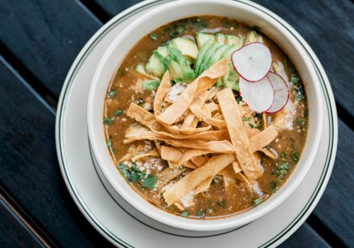 A bowl of soup topped with tortilla strips, sliced radishes, and avocado on a dark wooden table.