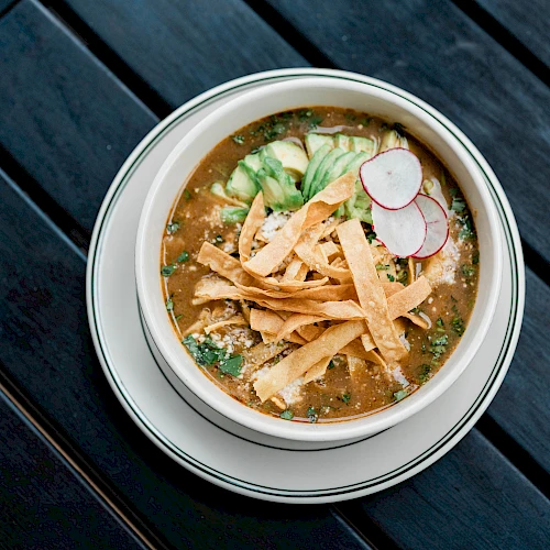 A bowl of soup topped with tortilla strips, sliced radishes, and avocado on a dark wooden table.