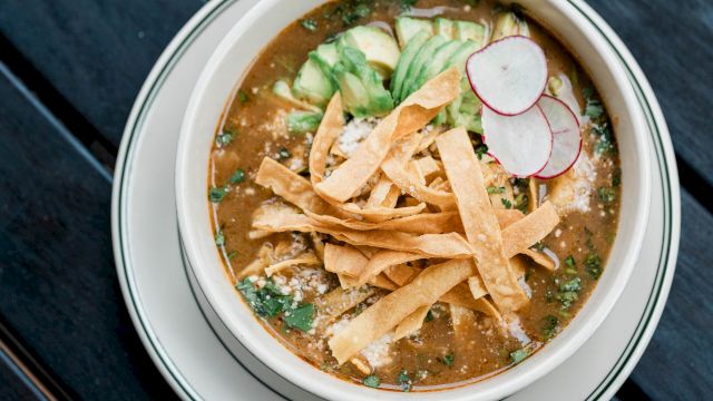 A bowl of soup topped with tortilla strips, sliced radishes, and avocado on a dark wooden table.