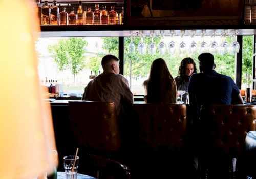 People sitting at a bar counter with hanging glasses and bottles above, viewed from behind. A lamp and drink in the foreground.