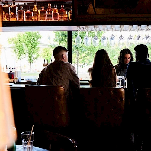 People sitting at a bar counter with hanging glasses and bottles above, viewed from behind. A lamp and drink in the foreground.