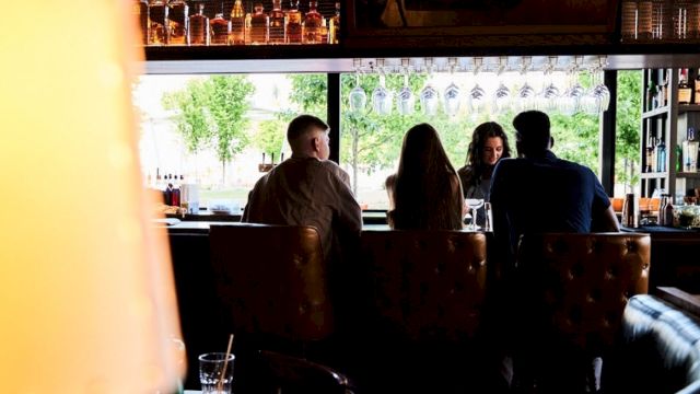 People sitting at a bar counter with hanging glasses and bottles above, viewed from behind. A lamp and drink in the foreground.
