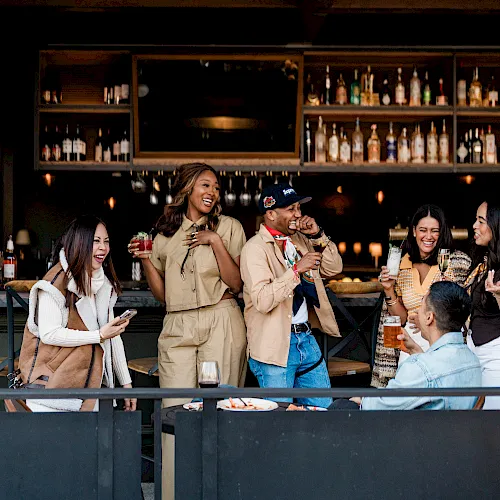 Friends laugh at a cozy bar, holding drinks and cocktails, under warm lights with shelves of bottles behind them.