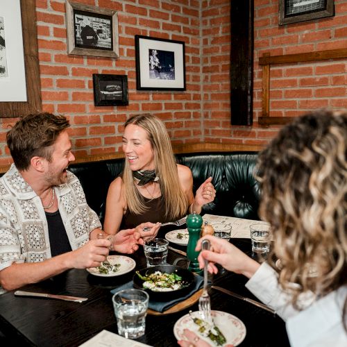 A group of people are smiling and eating together at a restaurant, with artwork on the brick wall behind them.