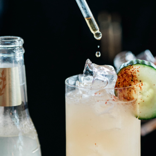 A bartender is crafting a cocktail with ice, cucumber garnish, and a dropper, next to a mixer bottle and shaker.