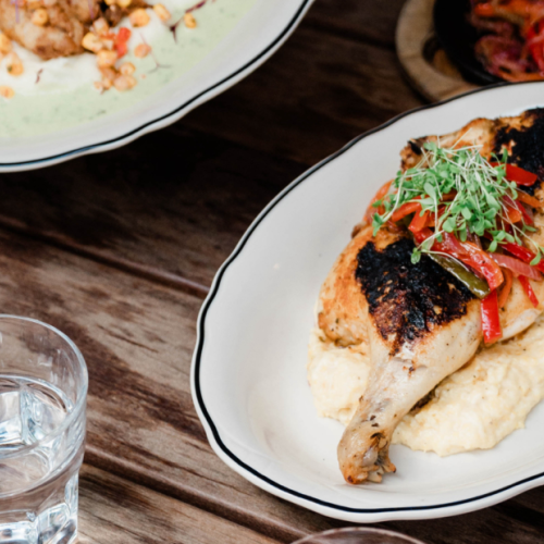 A wooden table with a glass of water and plates of roasted chicken on mashed potatoes and another creamy dish.