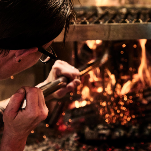 A person is tending a fire in a grill or oven, using a tool to adjust the logs or flames.