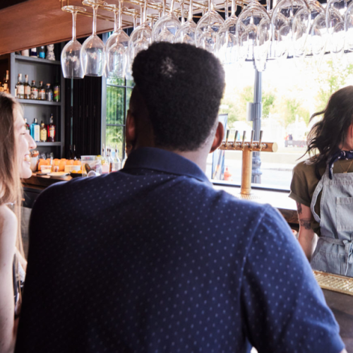 A bartender serves drinks while smiling at customers seated at a bar. Wine glasses hang overhead, with bottles visible on shelves.