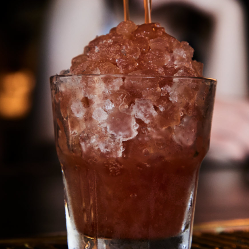 A glass filled with crushed ice and a reddish-brown liquid, possibly a cocktail, sits on a bar counter.