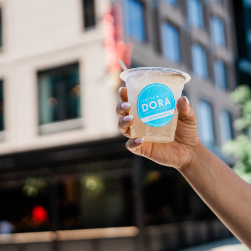 A person is holding a clear plastic cup with a straw and a blue "DORA" label on it, with buildings in the background.