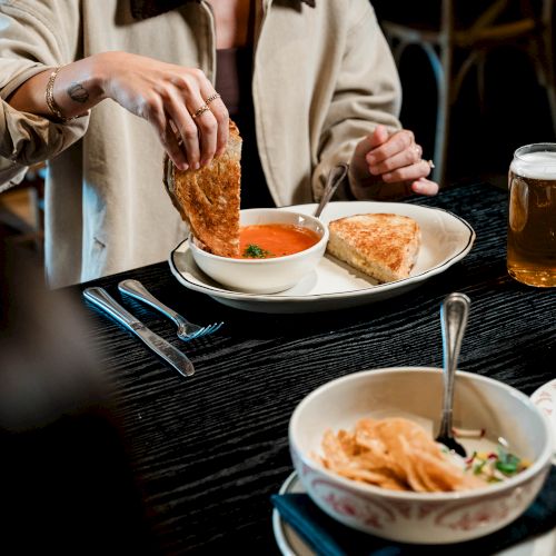 A person enjoys a meal with soup and a sandwich, accompanied by a bowl of chips and a glass of beer on a dark table.