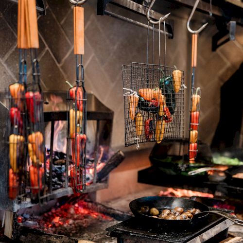 Vegetables being grilled over an open flame in a professional kitchen setting, with skewers and a pan nearby.