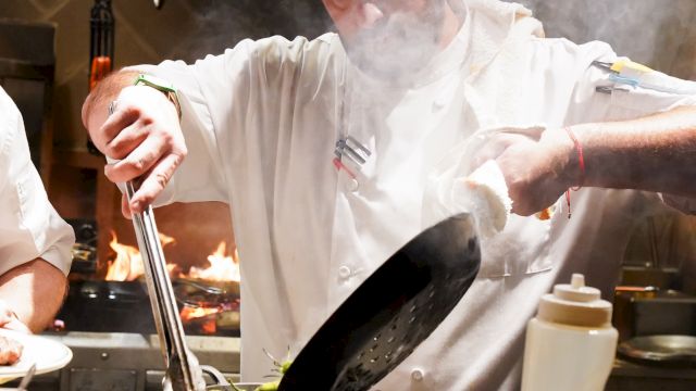 A chef is cooking, holding a pan and using tongs with steam rising, surrounded by kitchen utensils and ingredients on the counter.