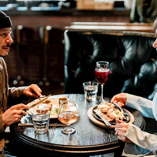 Two people are enjoying a meal together at a round table, with drinks and dishes in front of them, in a cozy setting.