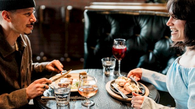 Two people are enjoying a meal together at a round table, with drinks and dishes in front of them, in a cozy setting.