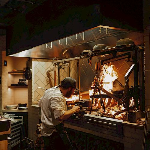 A chef grills over a blazing open flame in a busy kitchen, focused on flames and utensils under a hood.