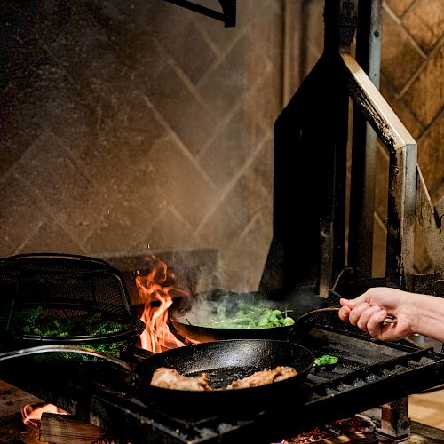 A person’s hand cooking on a stove with flames and food in a pan over an open fire, in a rustic kitchen.