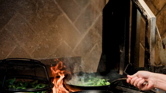 A person’s hand cooking on a stove with flames and food in a pan over an open fire, in a rustic kitchen.