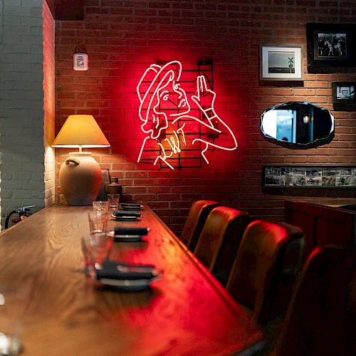 A cozy brick-walled bar with a neon red mural of a woman and a long wooden counter, warm lamp light, and framed art above the booth.