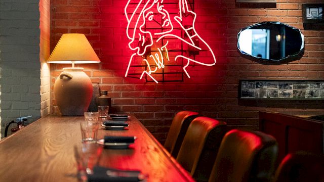 A cozy brick-walled bar with a neon red mural of a woman and a long wooden counter, warm lamp light, and framed art above the booth.