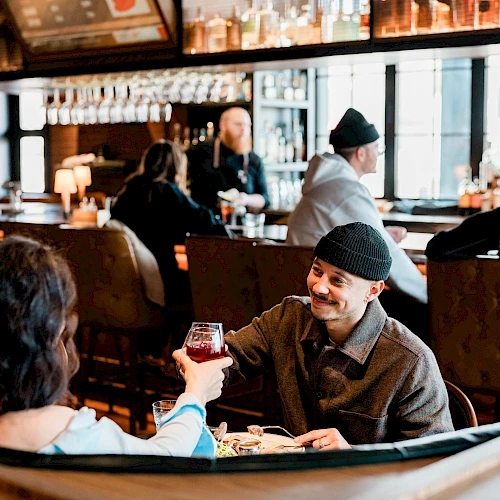 Two people share a toast over a meal in a cozy restaurant, smiling across a table with warm lighting and other patrons in the background.