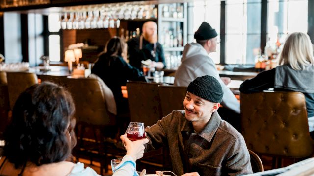 Two people share a toast over a meal in a cozy restaurant, smiling across a table with warm lighting and other patrons in the background.