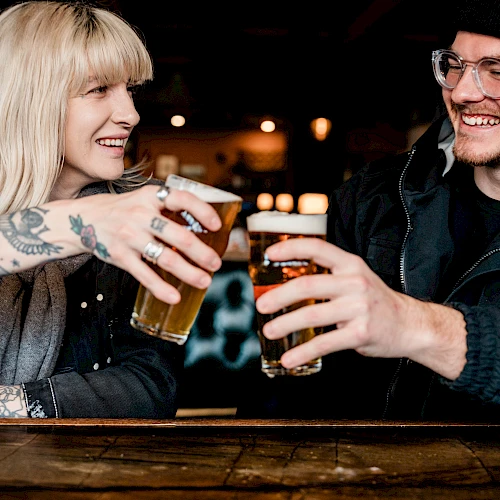 Two friends clink pint glasses at a bar, smiling and sharing a cheerful moment.