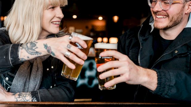 Two friends clink pint glasses at a bar, smiling and sharing a cheerful moment.