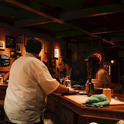 A cozy dimly lit bar scene with bartenders and patrons chatting at the counter, framed by bottles, glasses, and warm ambient lights.
