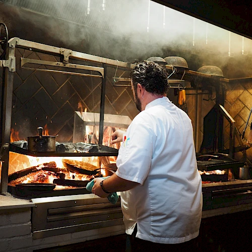 A chef in a white coat cooks on a sizzling grill in a smoky kitchen, flames and steam rise as food sizzles behind a glass shield.