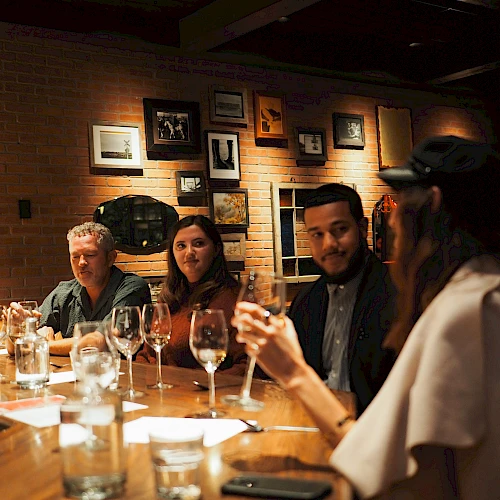 A small group sits around a wooden bar, drinking wine and chatting in a cozy, dimly lit restaurant with brick walls and framed photos.