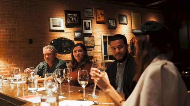 A small group sits around a wooden bar, drinking wine and chatting in a cozy, dimly lit restaurant with brick walls and framed photos.