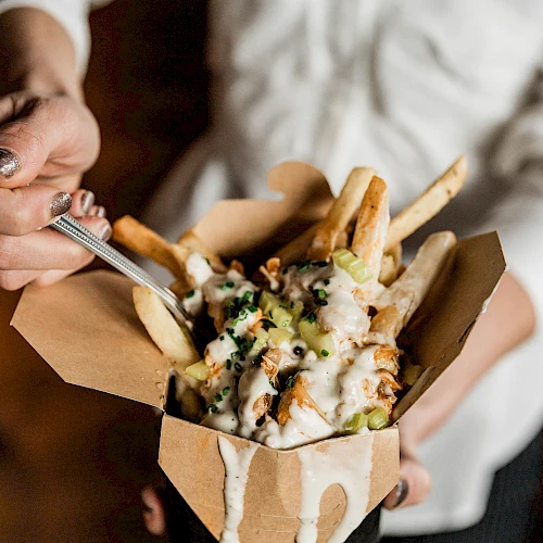 A person holds a takeout box with loaded fries, cheese, and sauce, drizzling as they use a fork to scoop some of it.