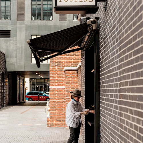 A person in a hat and light jacket stands at a doorway on a brick wall, near a sign that reads &ldquo;POTINE&rdquo; or &ldquo;POUTINE,&rdquo; in an urban alley.