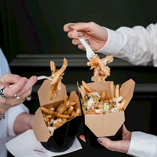 Two people sharing food: nachos and fries in brown boxes, dipping and grabbing crunchy toppings with chopstick-like hands, on a dark table.