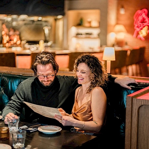 A couple sits in a dim, cozy restaurant booth, reading a menu together with smiles, blue-tinted lighting, and a stylish, intimate ambiance.
