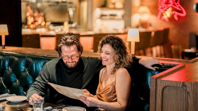 A couple sits in a dim, cozy restaurant booth, reading a menu together with smiles, blue-tinted lighting, and a stylish, intimate ambiance.