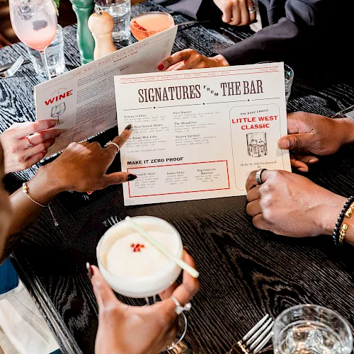 Two people sit at a bar, holding cocktail/wine menus labeled “Signatures FR THE BAR” and “Make it Zero Proof,” with drinks on the table.