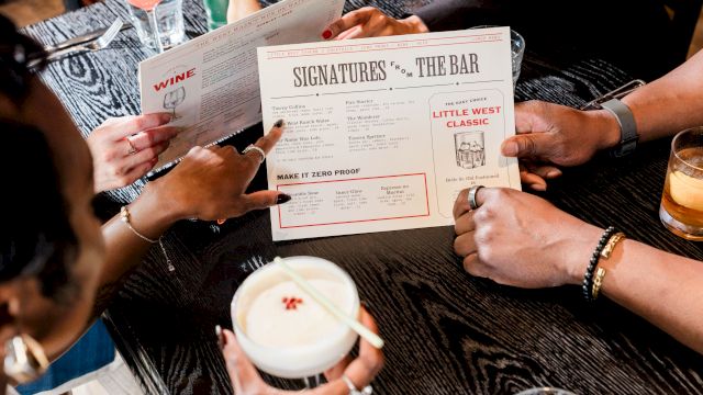 Two people sit at a bar, holding cocktail/wine menus labeled &ldquo;Signatures FR THE BAR&rdquo; and &ldquo;Make it Zero Proof,&rdquo; with drinks on the table.