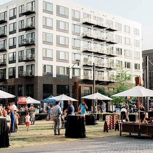 An outdoor gathering with people under white tents and a large apartment building in the background, on a sunny day.